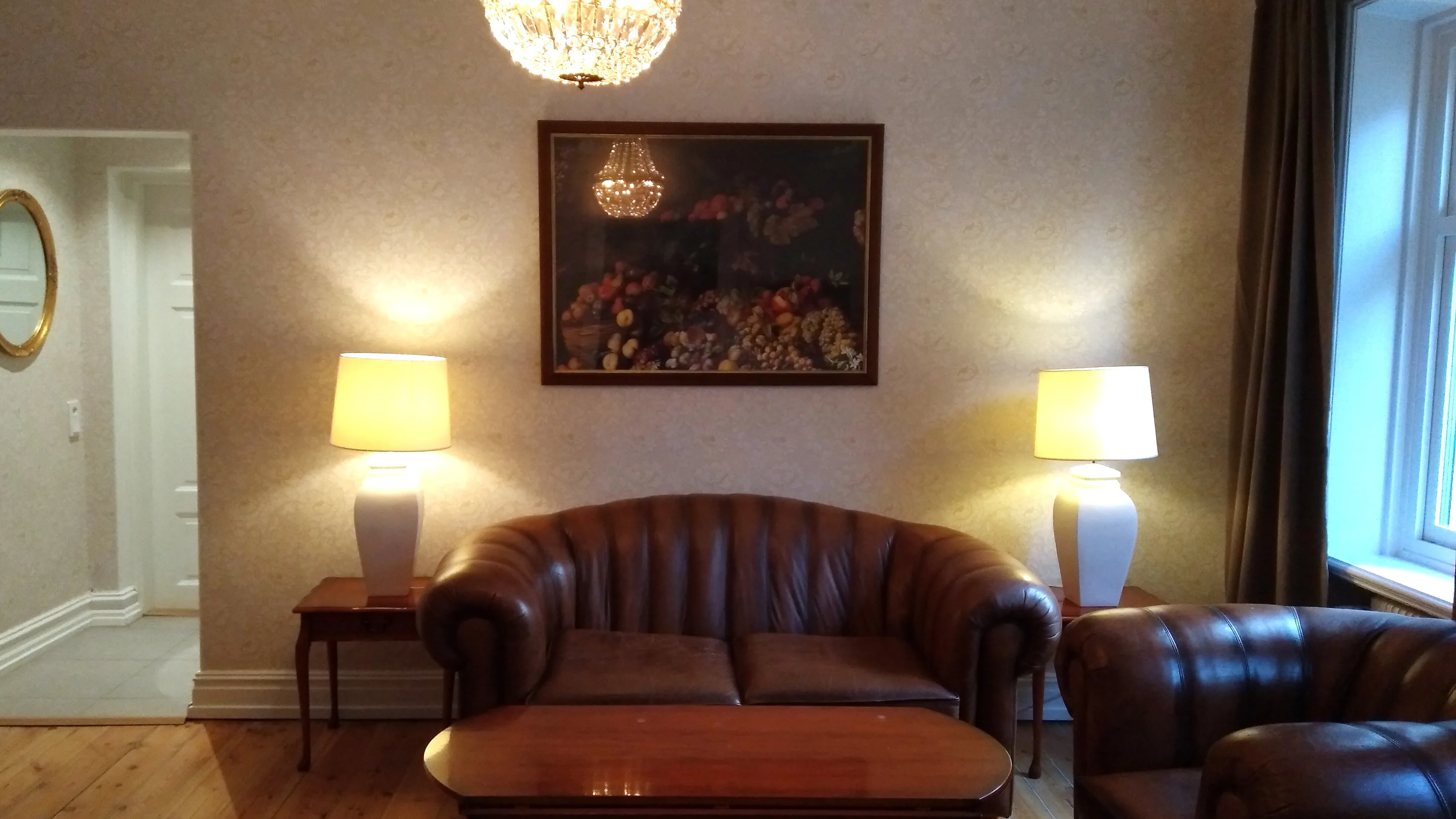 Ett hotellrum med ljusa, mönstrade tapeter och trägolv, med en kristallkrona i taket ovanför en brun soffgrupp i skinn.