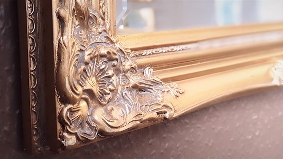 Kanten av en spegel med vackra gulddetaljer i snirkliga mönster, mot brunmålad vägg