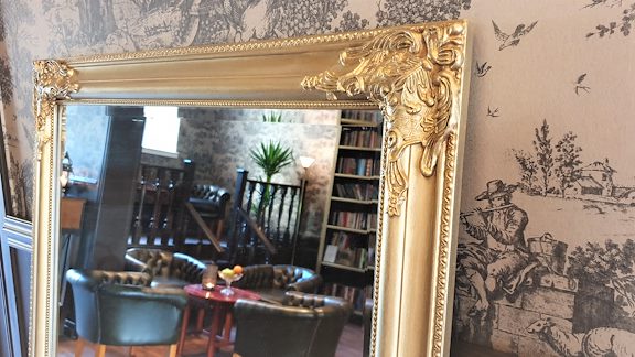 A mirror with gold details, with chairs and a bookshelf seen in the reflection.