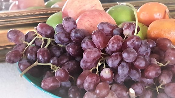 A bowl of fruit with purple grapes, green apples and oranges.