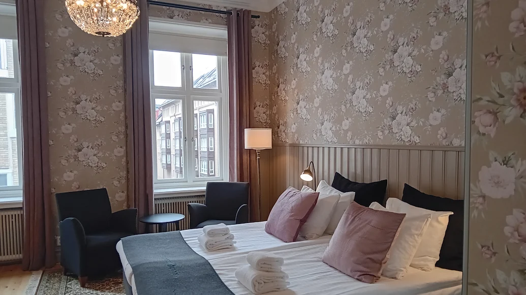 Ett dubbelrum med rosa blommiga tapeter och en kristallkrona i taket. Sängen har vita lakan och rosa prydnadskuddar, det står två fåtöljer framför fönstret.