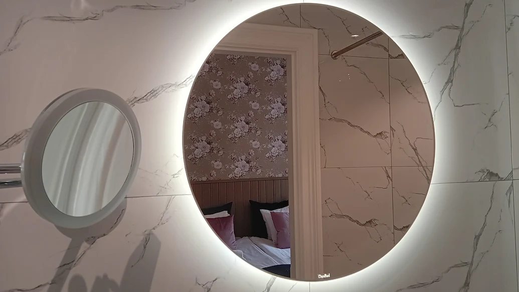 En stor, rund spegel upplyst bakifrån som speglar del av ett rum och badrummet den hänger i, väggarna har marmormönster.