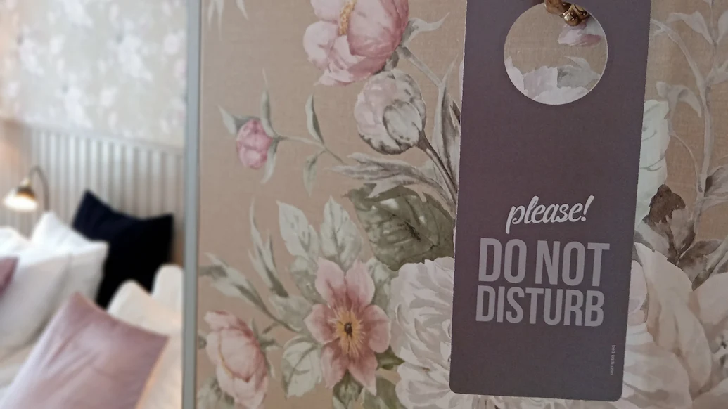En närbild på en Stör Ej-skylt, med en rosa tapet med blomstermotiv samt en säng i bakgrunden.