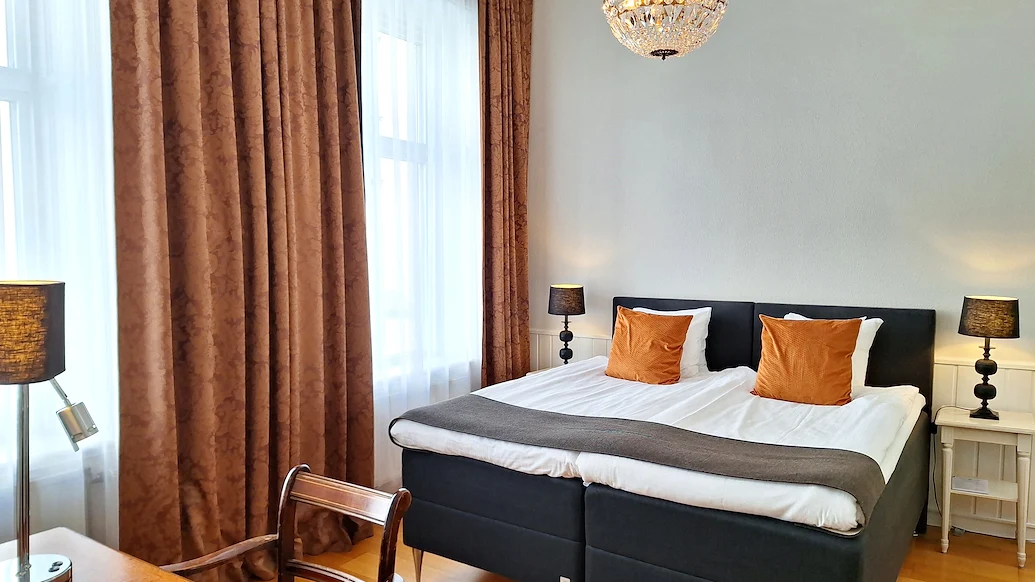 Ett dubbelrum med vita väggar, bruna gardiner och en kristallkrona i taket, och en dubbelsäng med vita lakan och orangea prydnadskuddar.