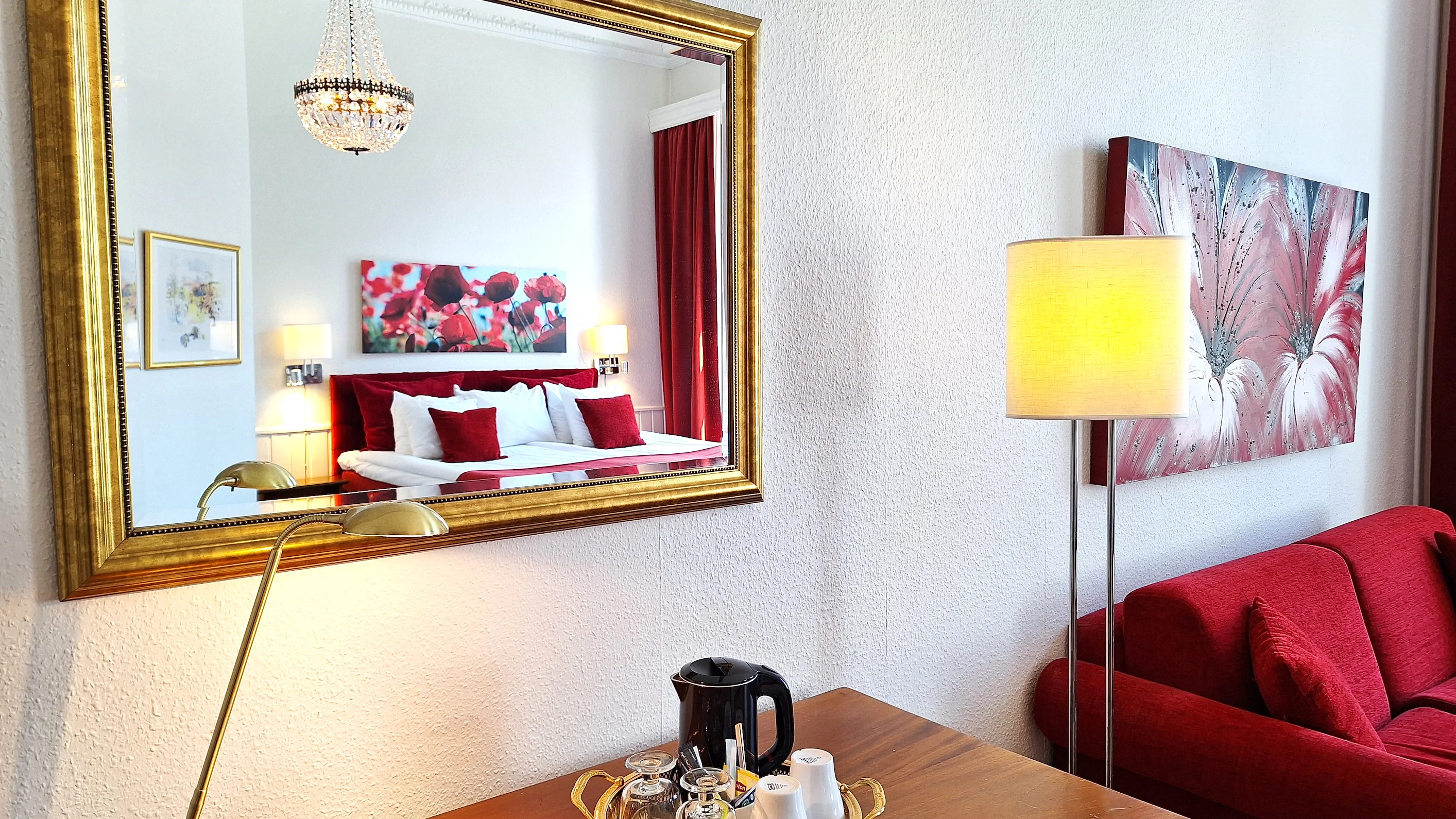 En spegel med guldram som hänger på en vit vägg, i spgelbilden ser man en kristallkrona, röd säng med vita lakan och röda prydnadskuddar. Bredvid spegeln står en röd soffa.
