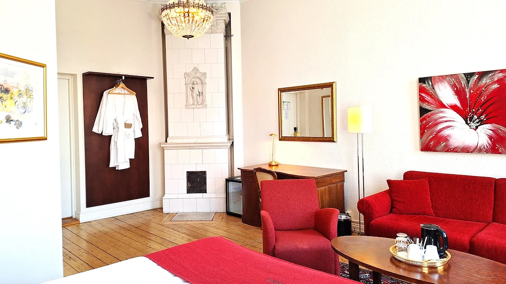 Ett ljust hotellrum med vita väggar, en kristallkrona i taken och en stor vit kakelugn. En röd soffgrupp står till höger om ett brunt skrivbord.