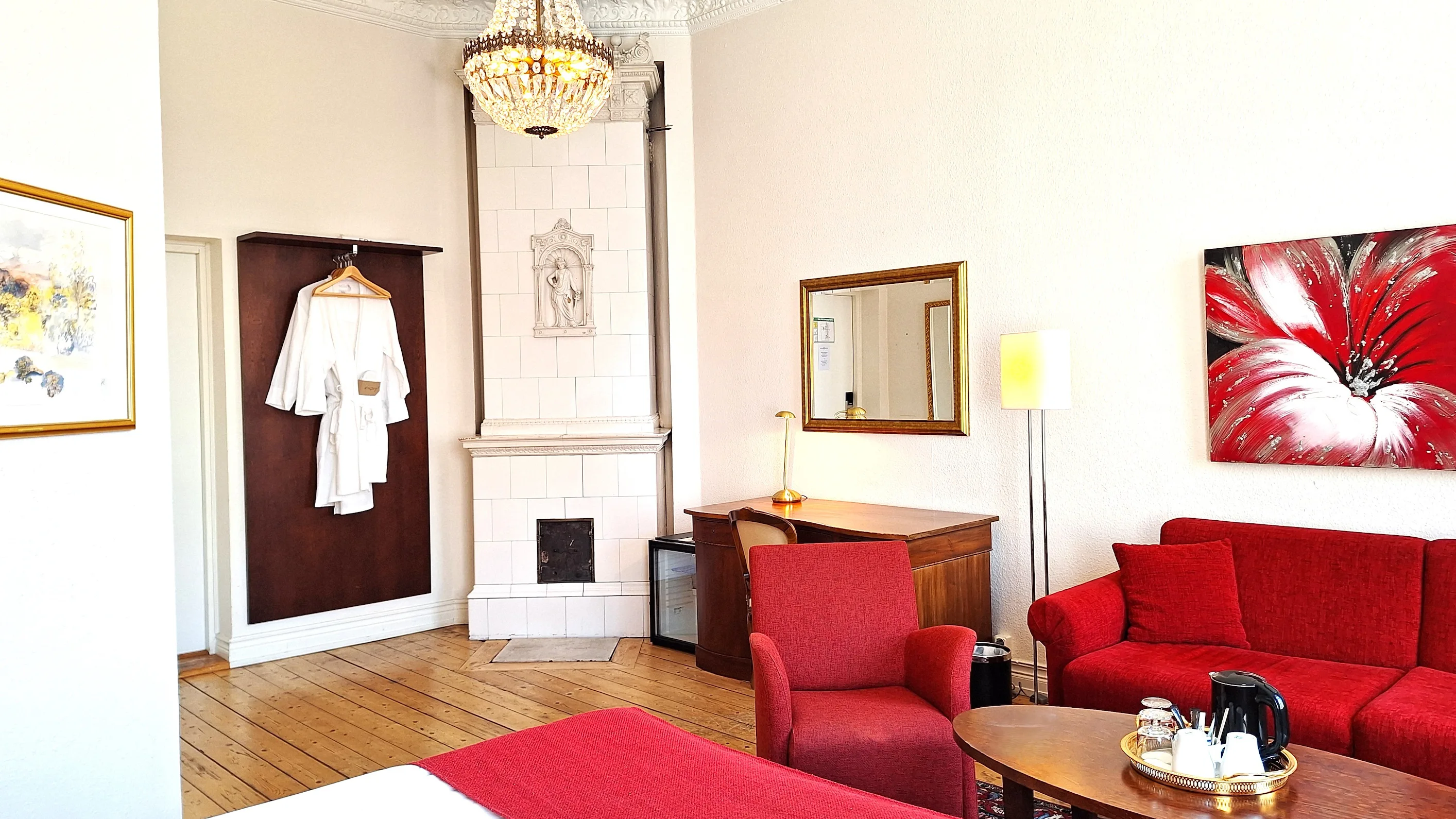 Ett ljust hotellrum med vita väggar, en kristallkrona i taken och en stor vit kakelugn. En röd soffgrupp står till höger om ett brunt skrivbord.