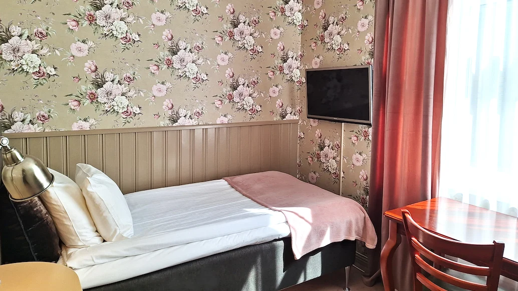 Ett hotellrum med blommig tapet, en säng med vita lakan framför en TV som hänger på väggen.