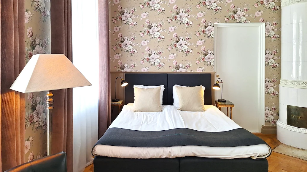 Dubbelrum med rosa blommiga tapeter, en hotellsäng och en vit kakelugn i hörnet.