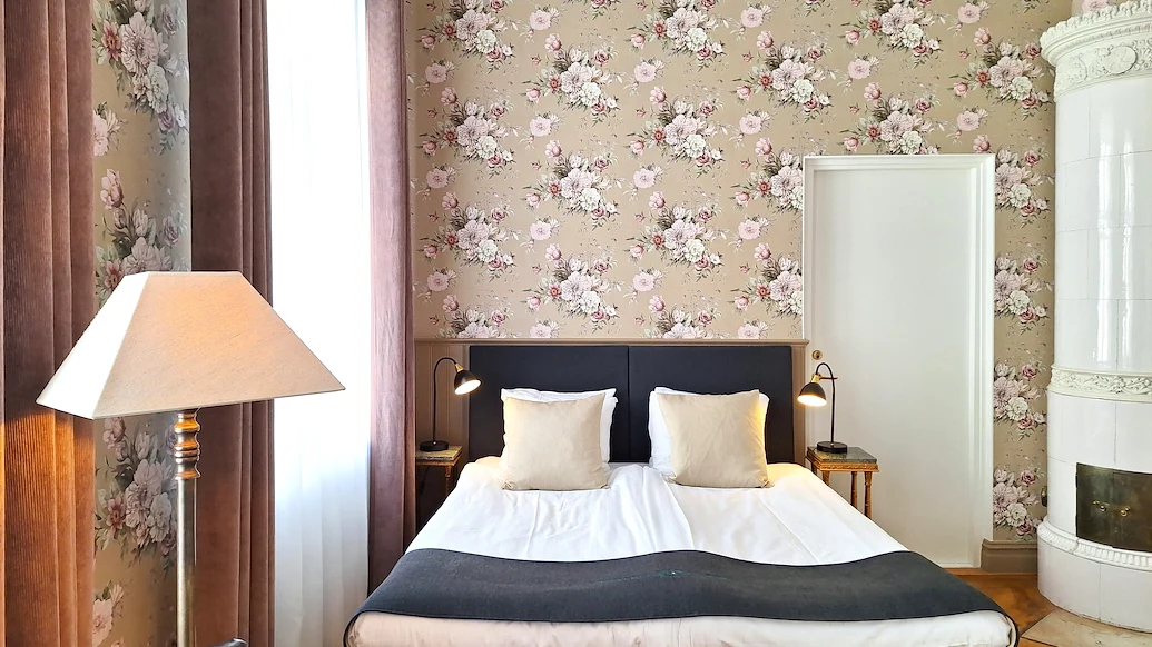 Dubbelrum med rosa blommiga tapeter, en hotellsäng och en vit kakelugn i hörnet.