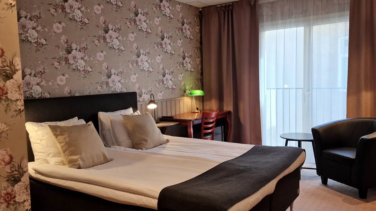 En dubbelsäng i ett rum med blommiga tapeter, en skinnfåtölj och en fransk balkong.