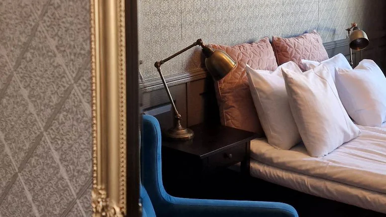 En närbild på en spegel med guldram, som speglar en säng med vita lakan och huvudkuddar, samt stora rosa prydnadskuddar.