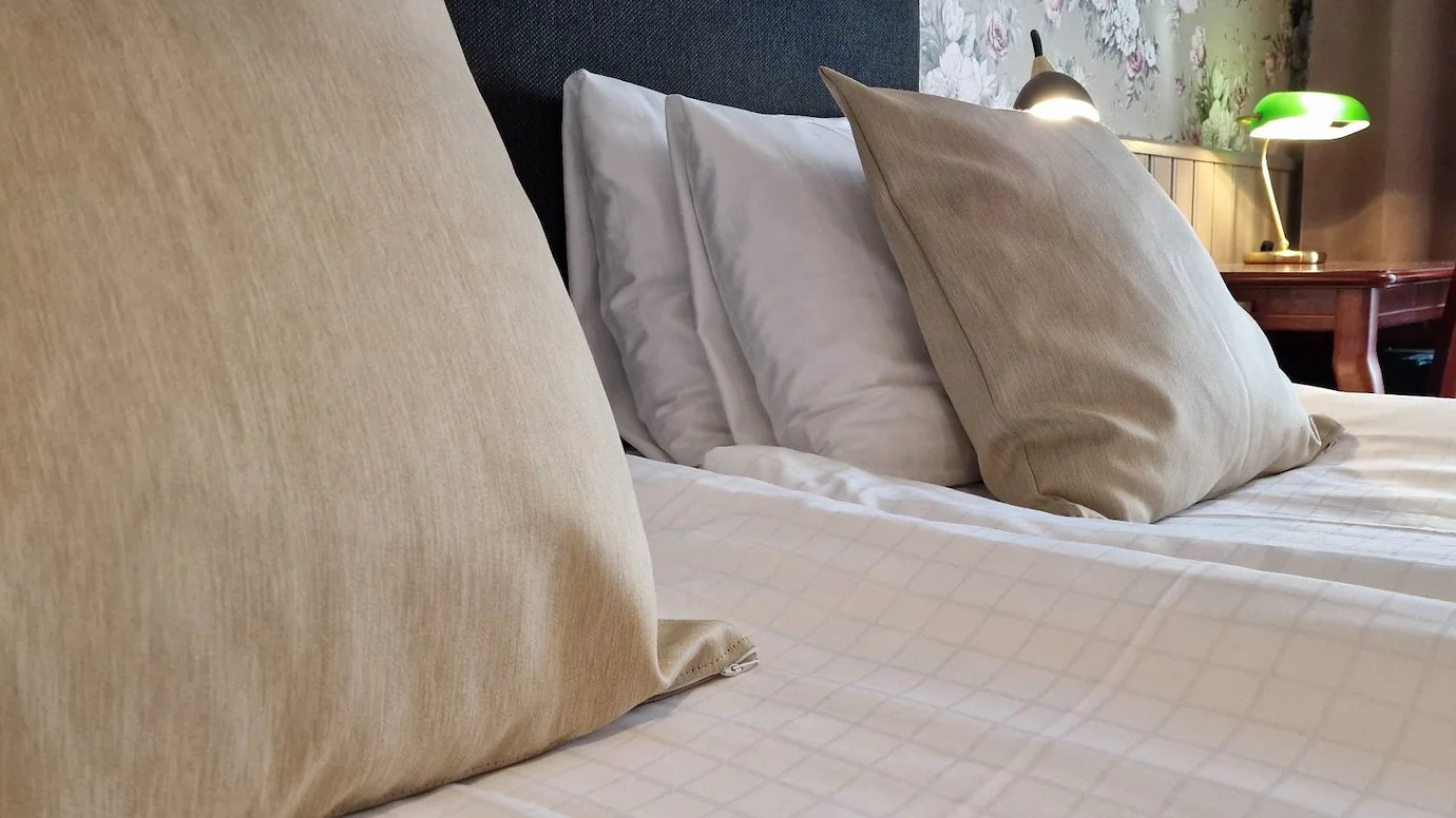 Närbild på en säng med vita lakan, vita huvudkuddar och två beiga prydnadskuddar.