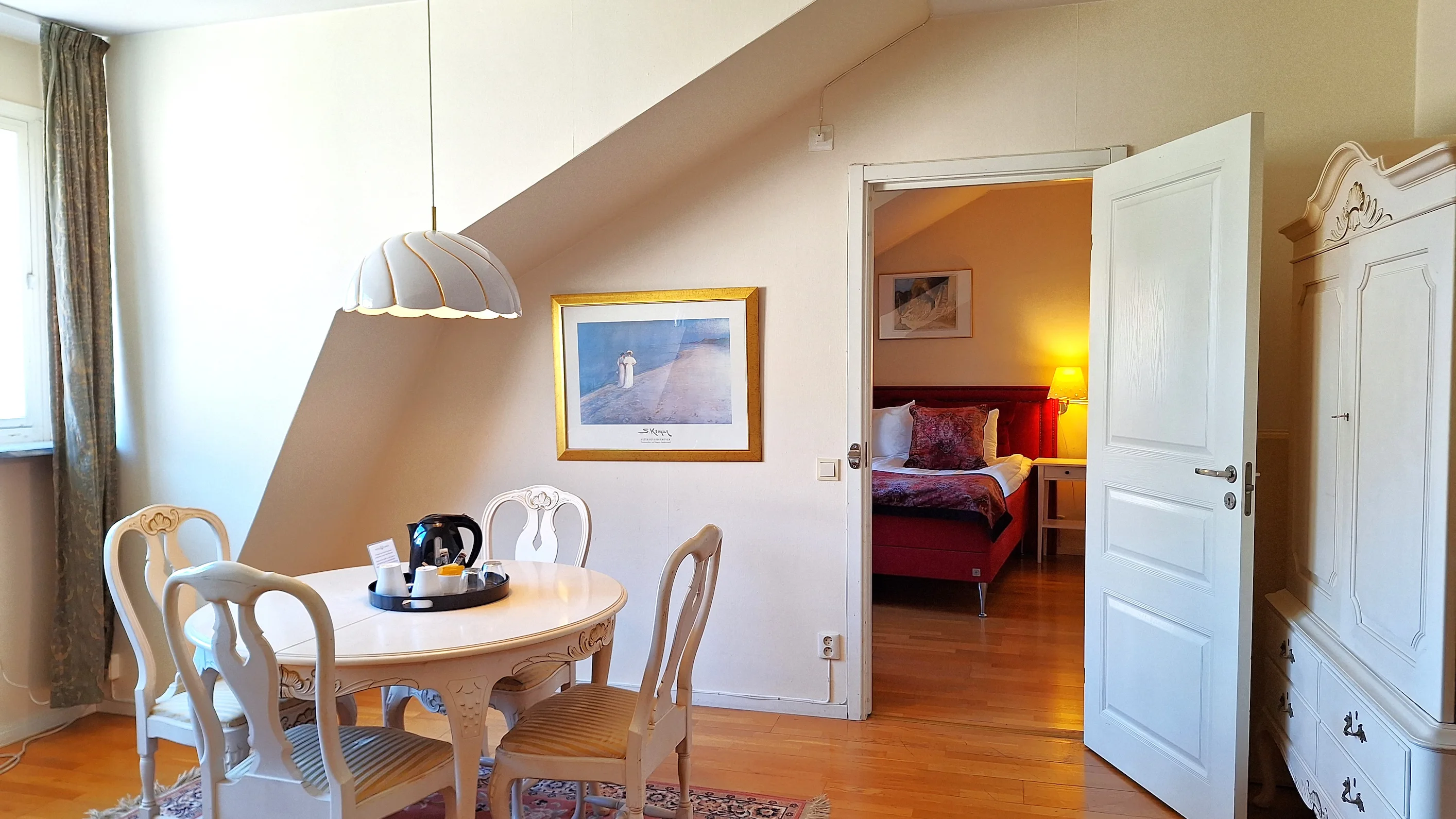 Ett hotellrum med ljusgula väggar, en vit matgrupp med fyra stolar, och en öppen dörr in till sovrums-delen av hotellrummet.