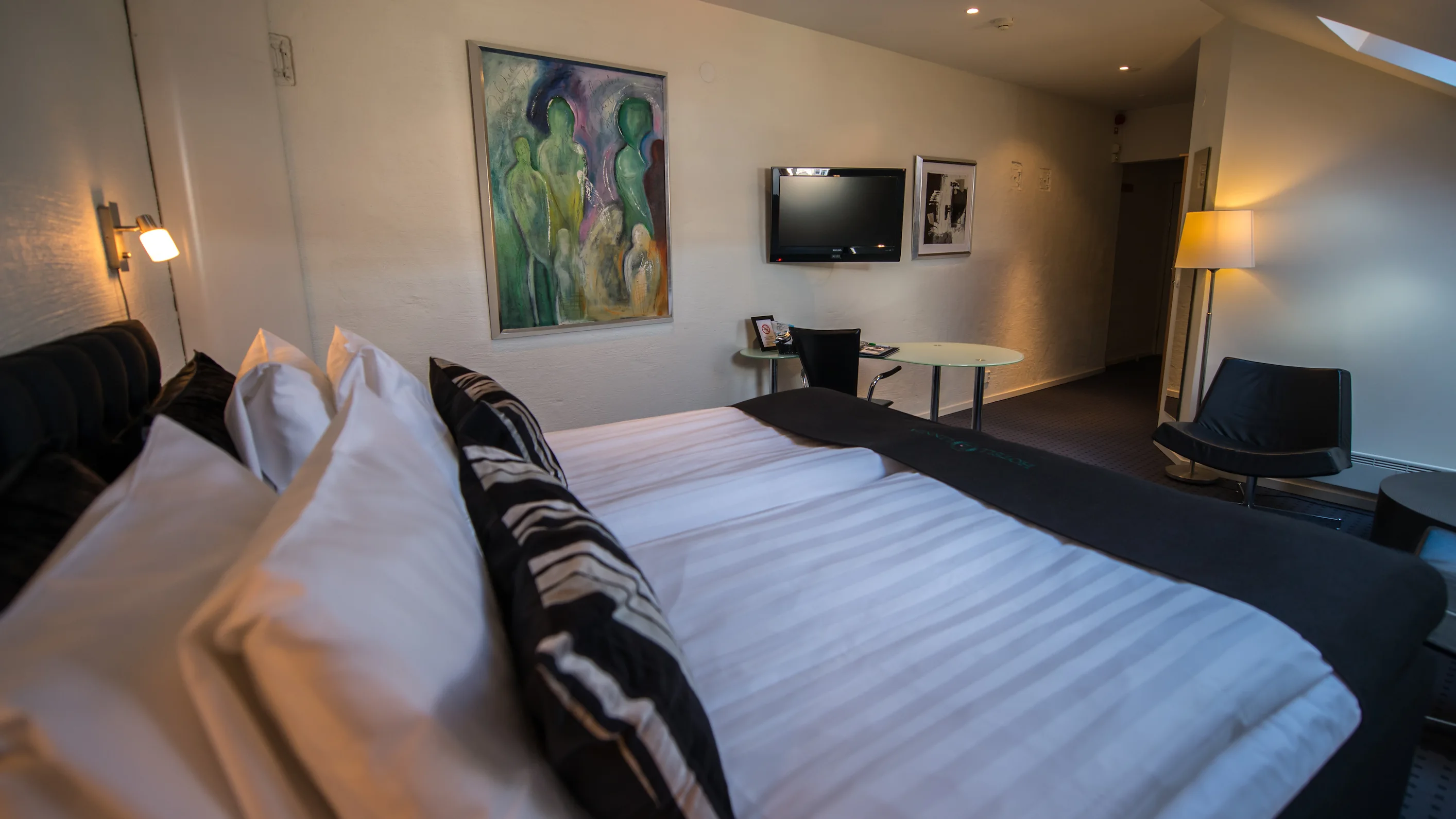 Hotellrum med vita väggar och snedtak, en stor säng i förgrunden med vita lakan och mörkt överkast, och ett skrivbord under en TV på väggen.