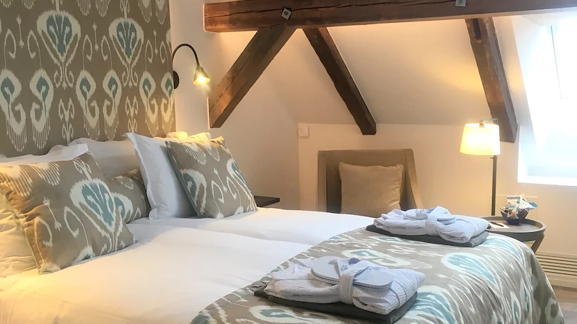 Ett ljus hotellrum med mörka, synliga träbjälkar och snedtak bakom en vit hotellsäng med ljusbruna prydnadskuddar.