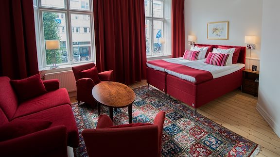 A spacious room with white walls, a red double bed and a red sofa and matching armchairs.
