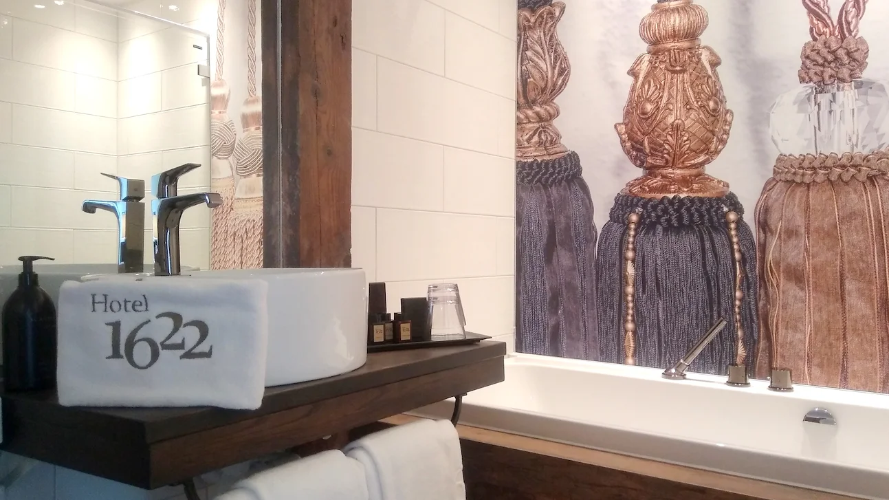 Ett handfat och badkar inuti ett badrum, med en handduk där det står Hotel 1622 hängandes från handfatet.