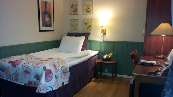 A room with a single bed with white sheets and a desk