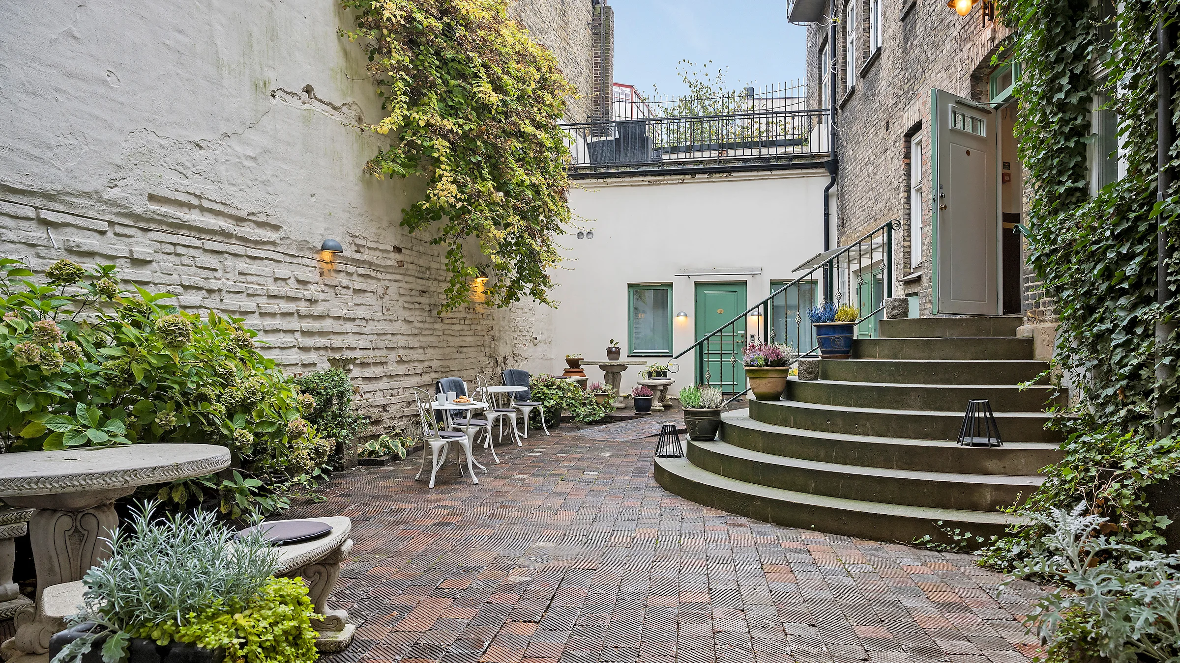 A courtyard with brick walls covered by shrubs and ivy. There`s a number of seating areas, and a green door to a hotel room.