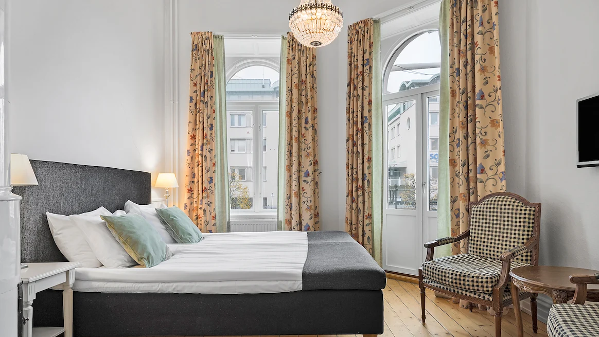 Ett hotellrum med vita väggar, gula gardiner och en kristallkrona. En stor dörr leder ut till en balkong, mitt emot står en grå säng med vita lakan.