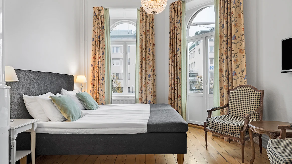 Ett hotellrum med vita väggar, gula gardiner och en kristallkrona. En stor dörr leder ut till en balkong, mitt emot står en grå säng med vita lakan.