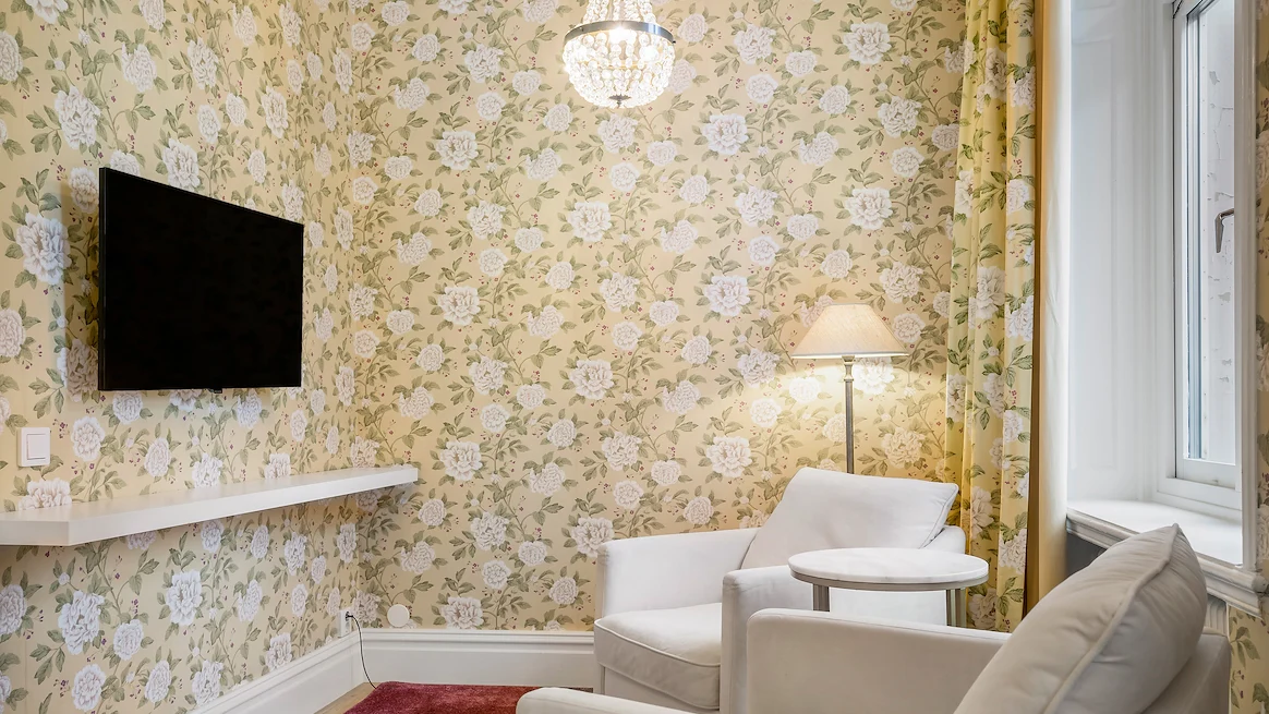 Ett hotellrum med gula tapeter med vita blommor, en TV på väggen och två ljusa fåtöljer framför.
