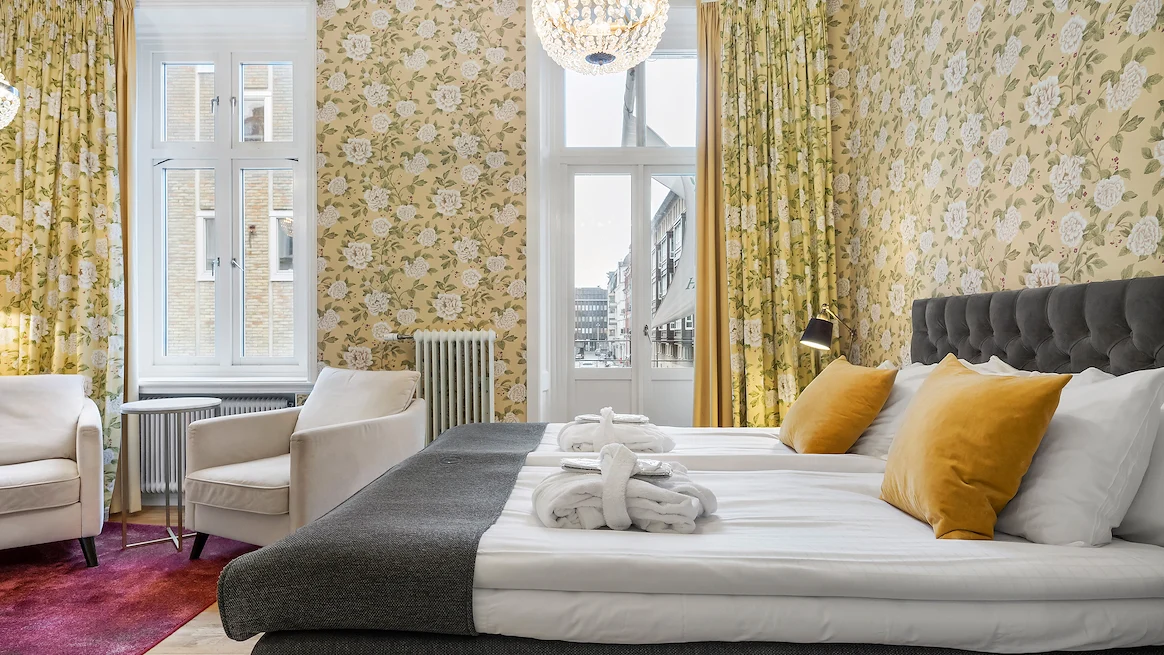 Ett hotellrum med gula tapeter, kristallkrona i taket och två vita fåtöljer till vänster om en stor mörkgrå säng med vita lakan. En dörr leder ut till en balkong.