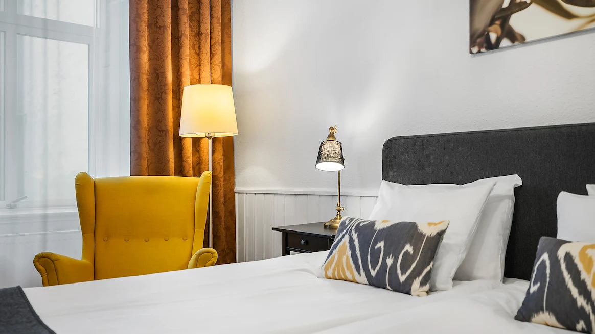 Ett ljust hotellrum med en gul fåtölj vid fönstret, samt en dubbelsäng med vita lakan och blågula prydnadskuddar.