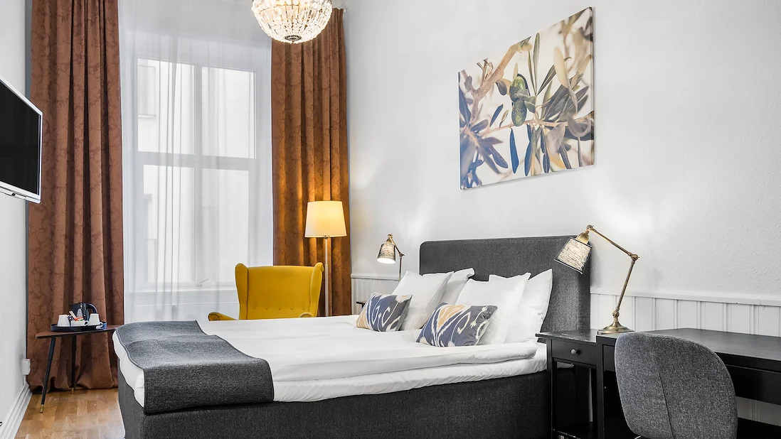 Ett ljust dubbelrum med vita väggar, kristallkrona i taket och en grå säng med vita lakan.