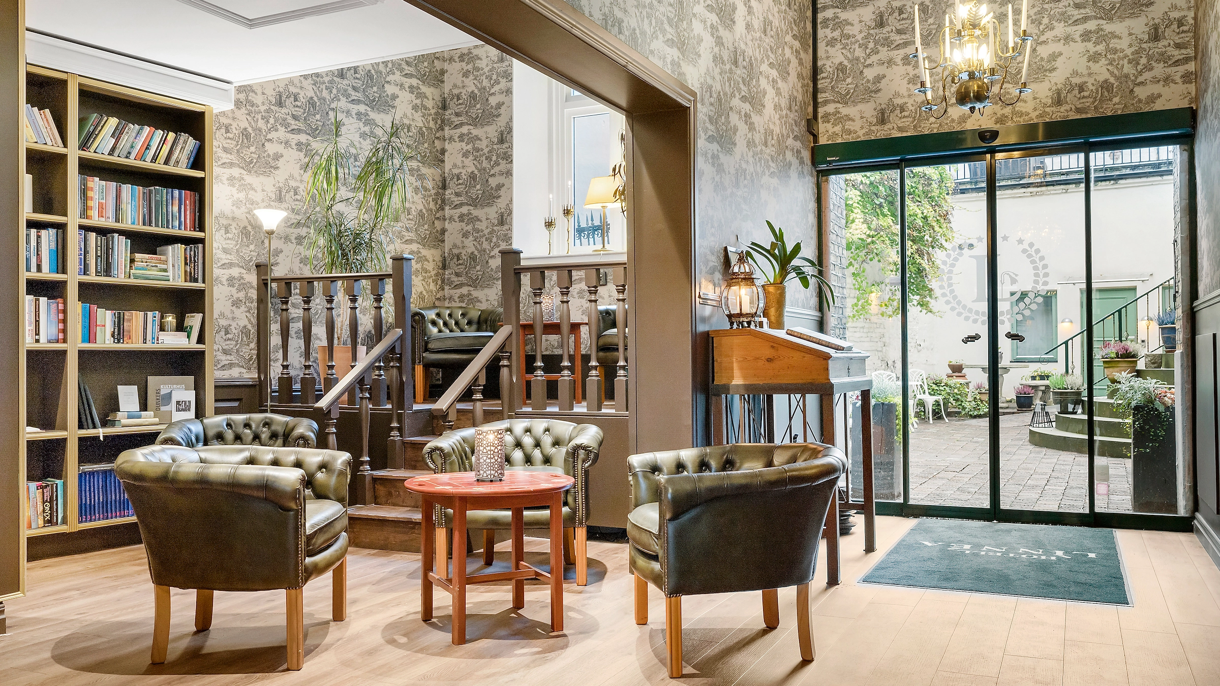 Hotellets lobby och bibliotek, med bokhylla till vänster om tre gröna skinnfåtöljer runt ett bord, med glasdörrar till hotellets innergård till höger.