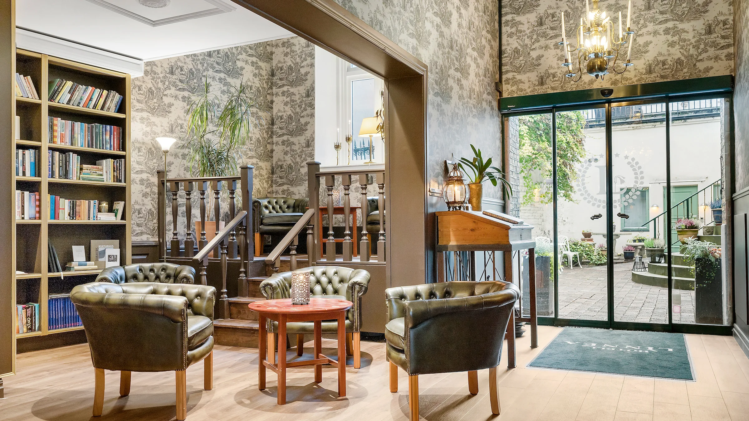 A lobby and library, with a bookcase to the left of three green leather chairs around a table. To the right glass doors leads to the hotel courtyard.