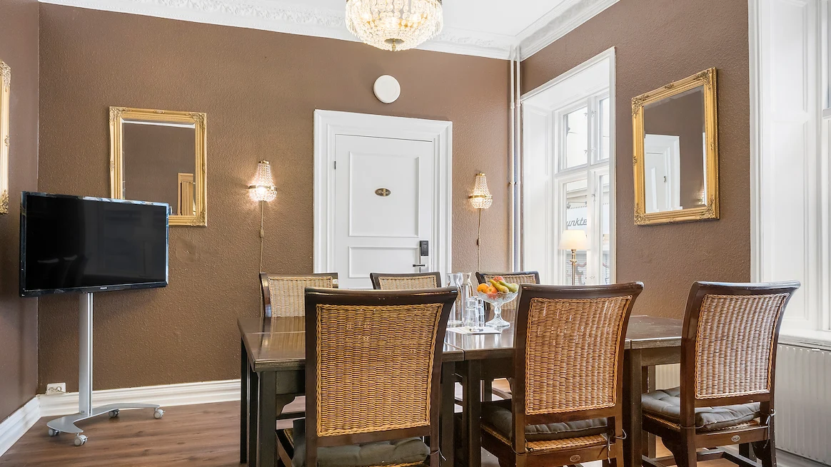 Ett mötesrum med bruna tapeter och kristallkrona i taket, med bord och stolar uppställda framför en TV-skärm.
