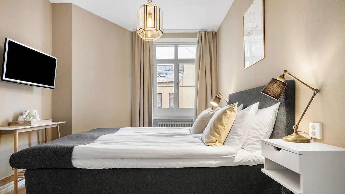 Ett ljust, mindre hotellrum med beiga väggar, mörkgrå säng med vita lakan och beiga prydnadskuddar.