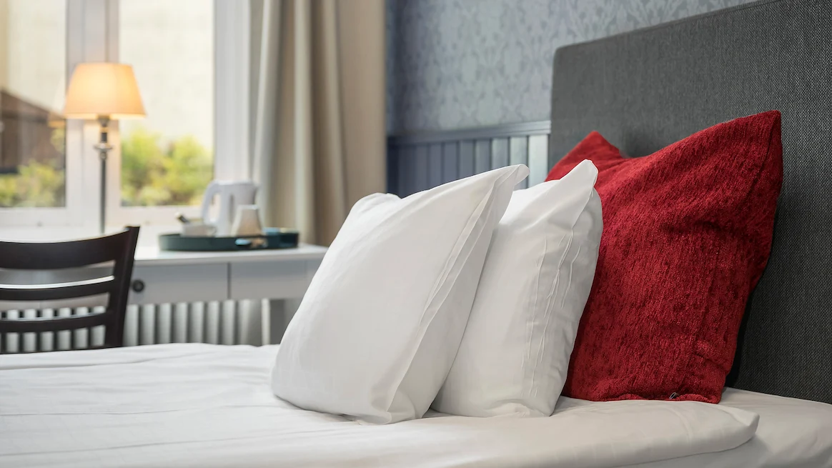 A close up shot of two white pillows leaning against a red decorative pillow on a single bed in front of a window.