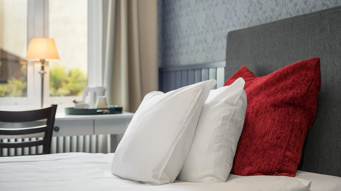 A close up shot of two white pillows leaning against a red decorative pillow on a single bed in front of a window.