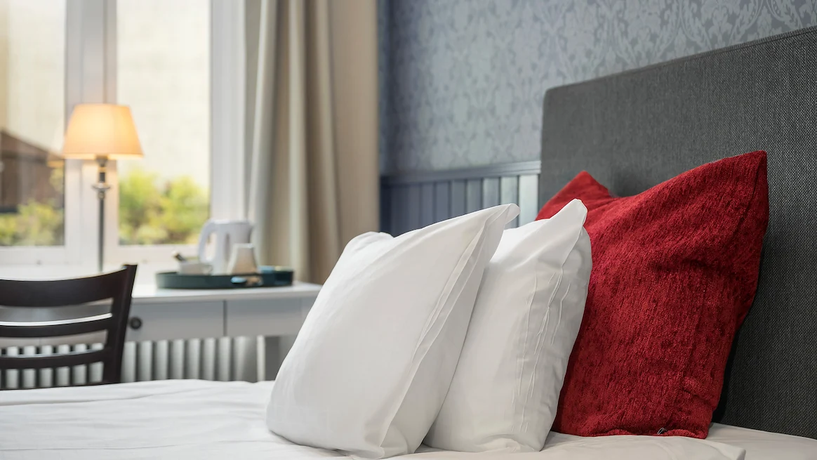 Närbild på en säng med två vita huvudkuddar som lutar mot en stor, röd prydnadskudde, framför ett fönster i ett ljus hotellrum med blå tapeter.