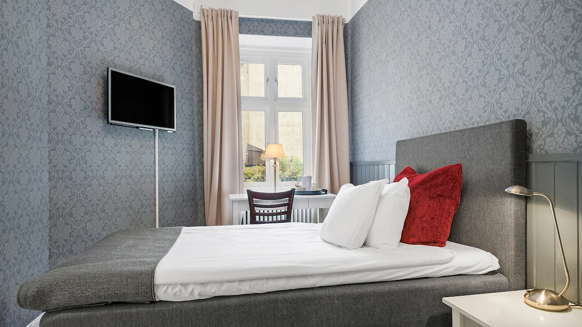 Ett hotellrum med blå mönstrade tapeter, en grå Enkelsäng med vita lakan och en TV som hänger på väggen mitt emot.