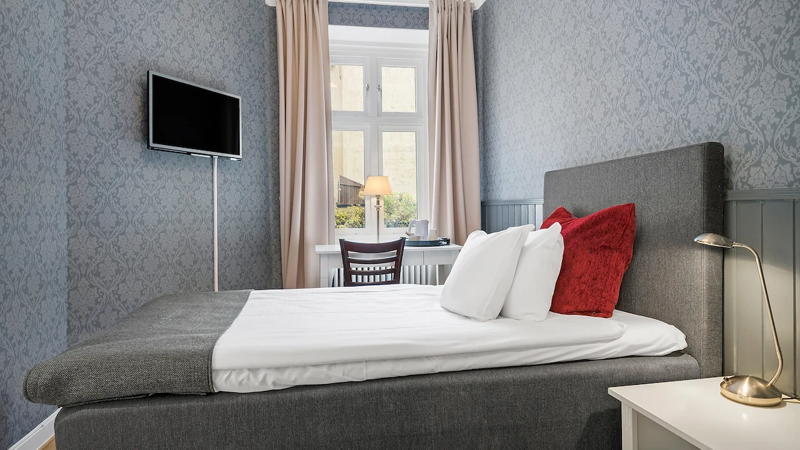 A room with blue patterened wallpapers, a single bed in front of a wall-mounted TV and a small desk in front of a window.