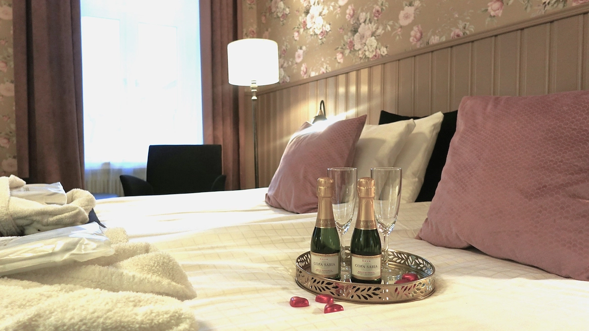 Ett fat med två små flaskor mousserande vin och två champagneglas står på en dubbelsäng med rosa prydnadskuddar.