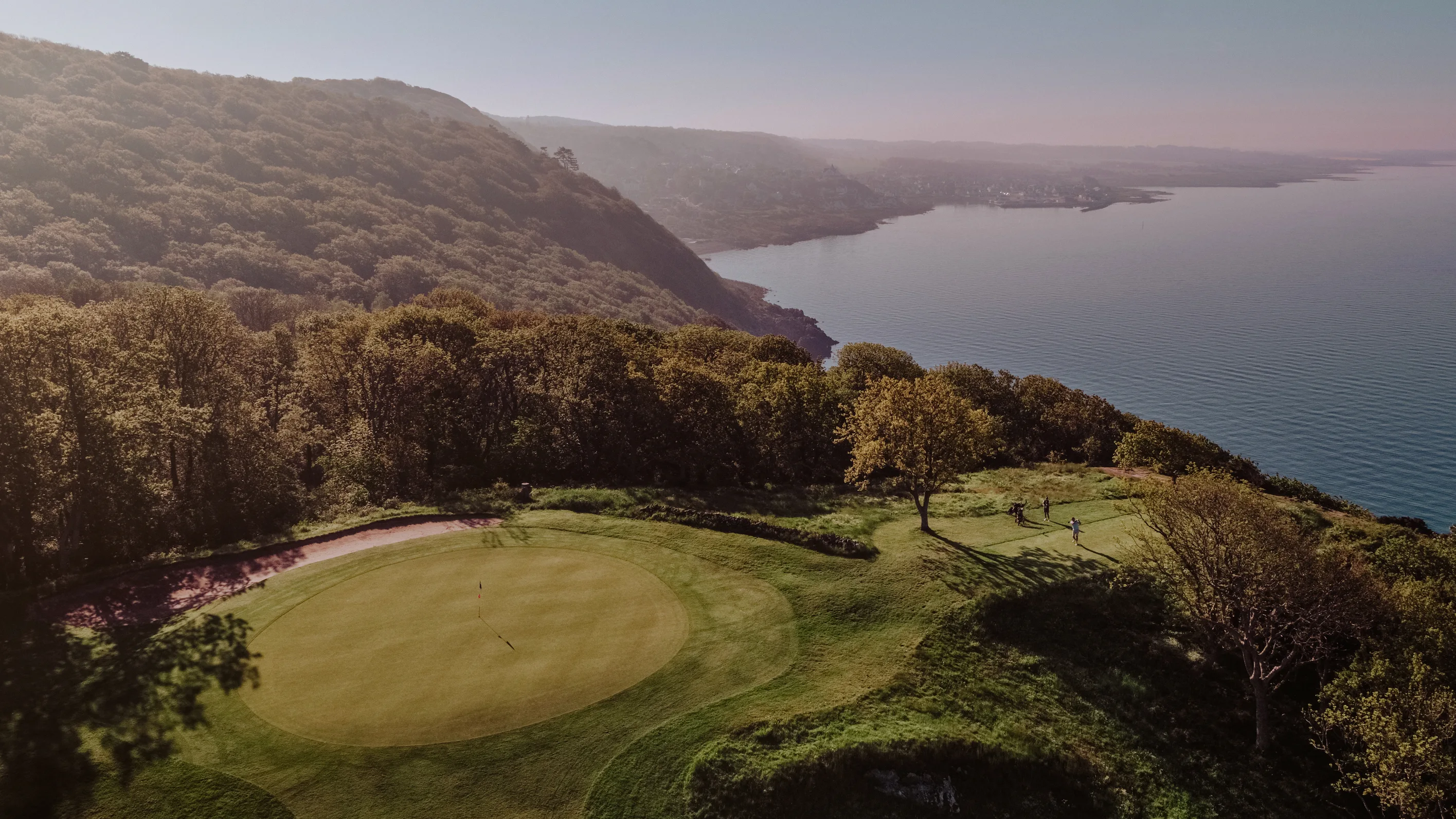 En golfbana på en kulle ovanför havet omringat av skog, en solig dag.