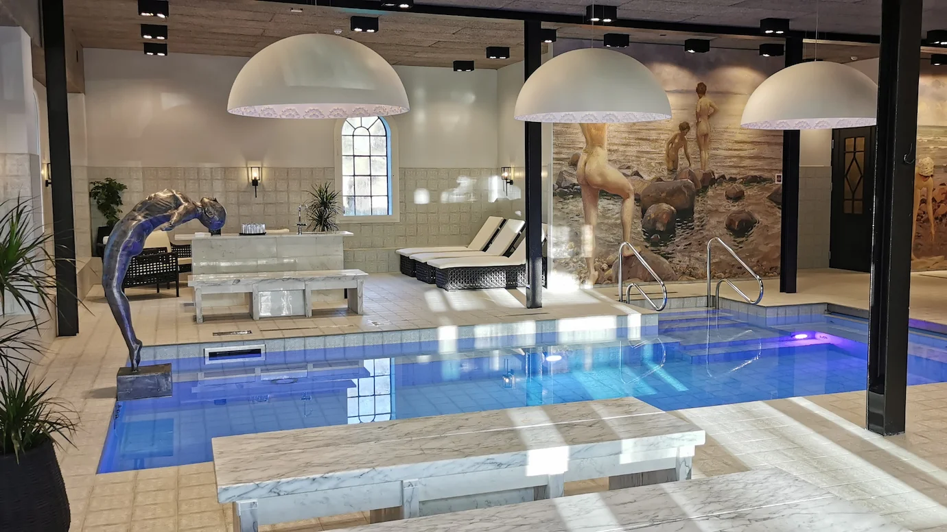 Ett rum med vitt kakel och en stor simbassäng i mitten, med en staty av en kvinna som dyker baklänges i ena änden.