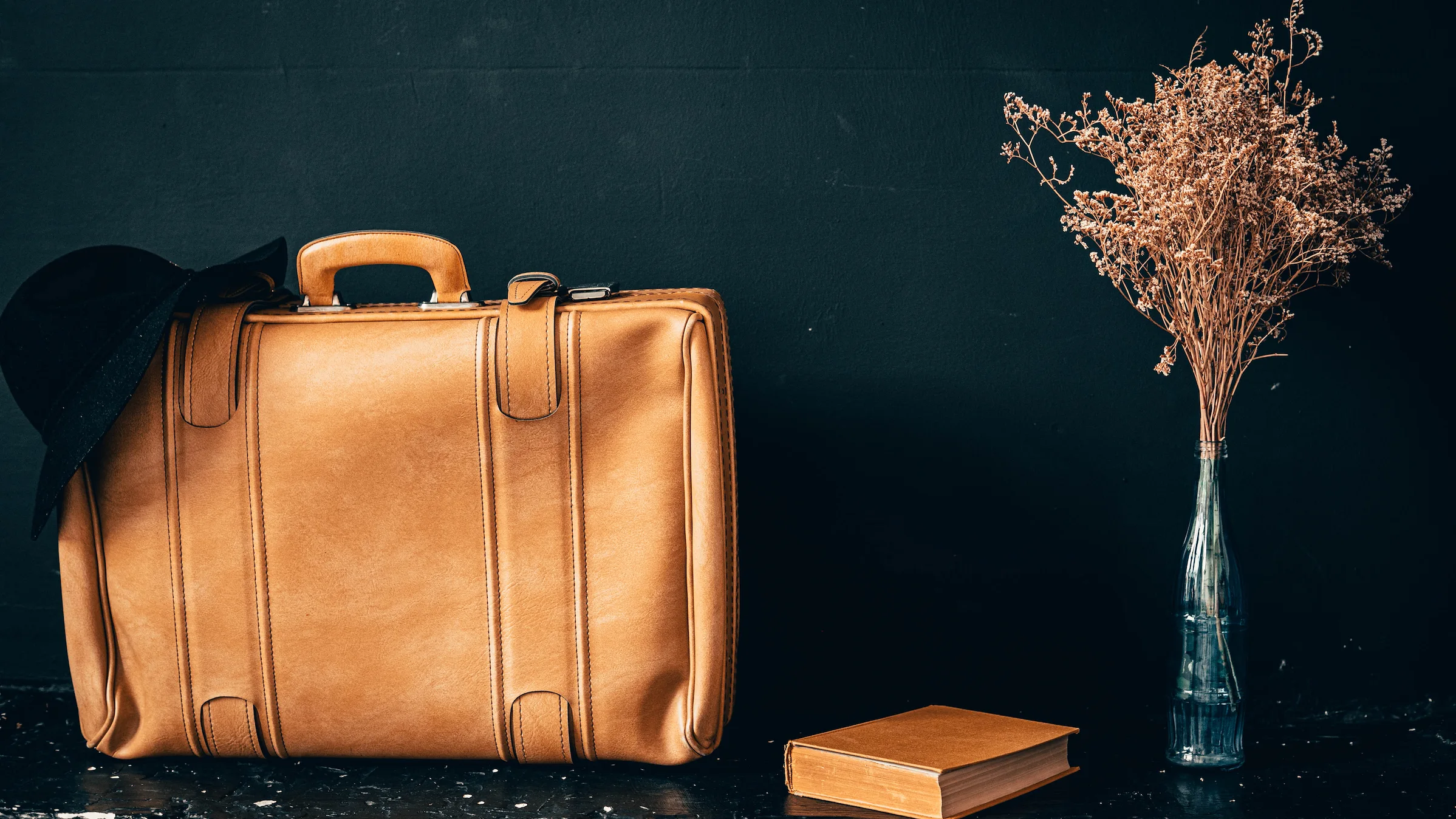 A hat resting on a light brown leather bag, a book and a vase with dried flowers against a black background.